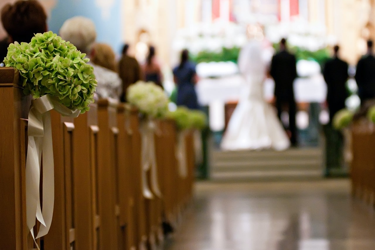 Close up of floral arrangements on church pews at a wedding with bride and groom in the background