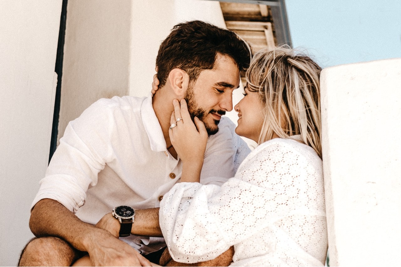 A couple sitting close together about to kiss. The woman has her hand on the man’s face wearing an engagement ring