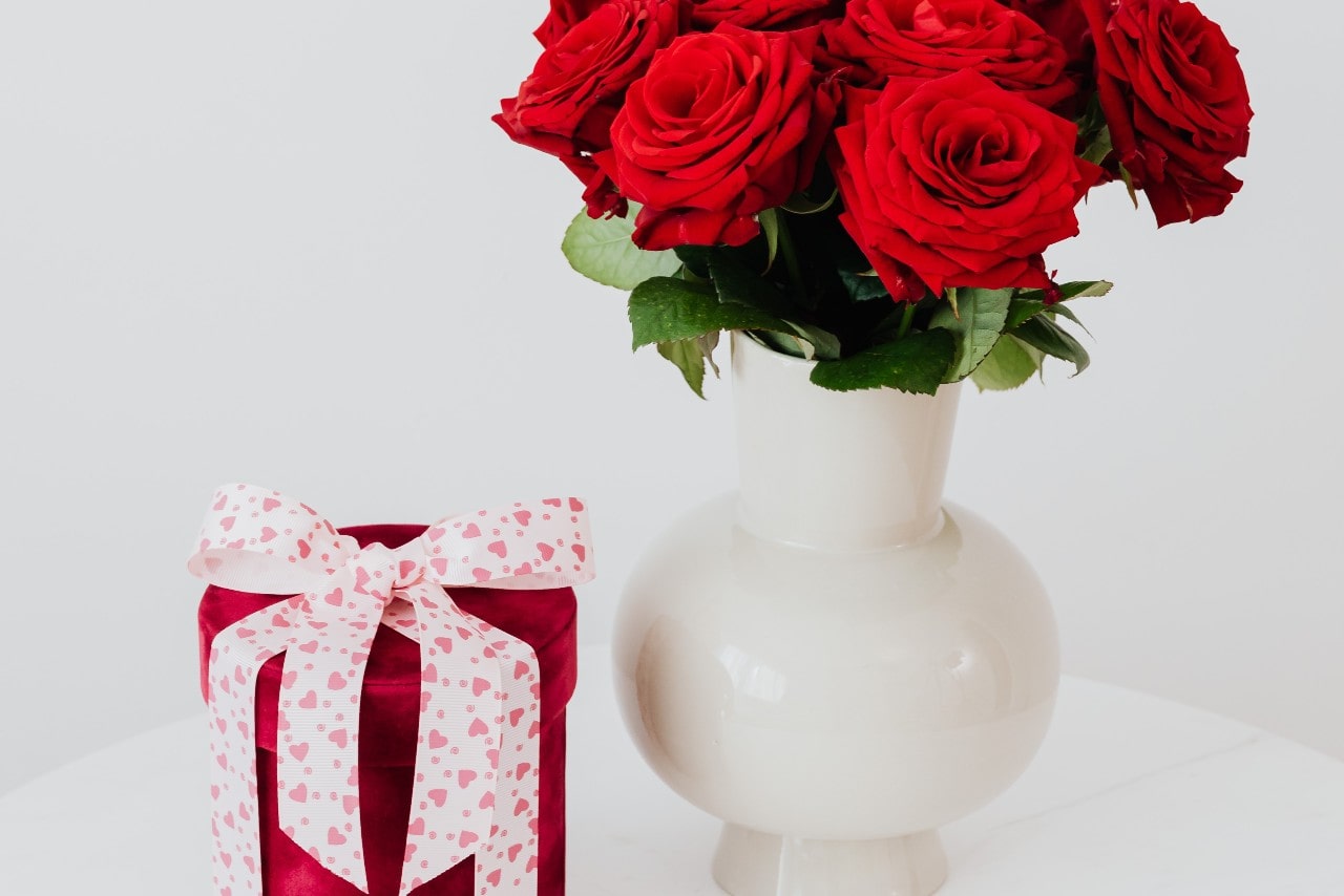 A velvet, cylinder gift box with a white and pink hearts bow on top next to a white vase with a bouquet of red roses