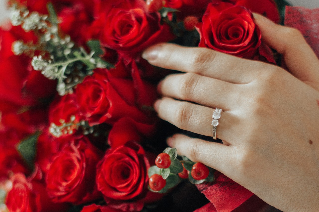 A hand with a three stone diamond engagement ring is resting on a bouquet of red roses and baby breath