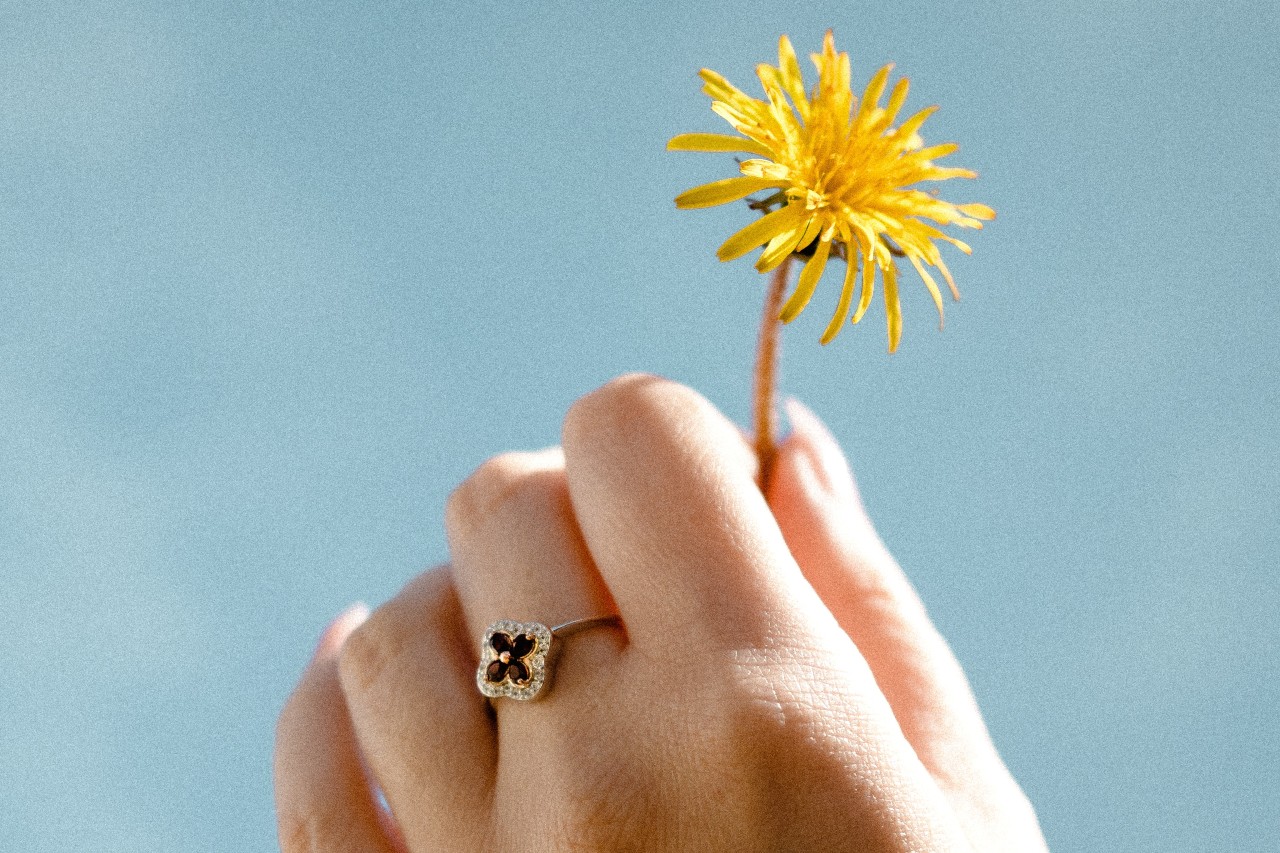 A hand holding a dandelion against the sky and wearing a clover-inspired fashion ring