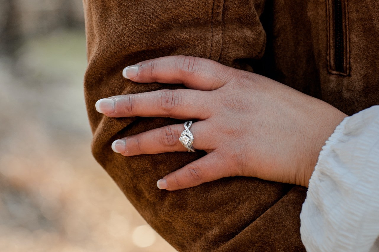 A woman’s hand resting on someone’s jacketed arm, wearing a princess cut engagement ring