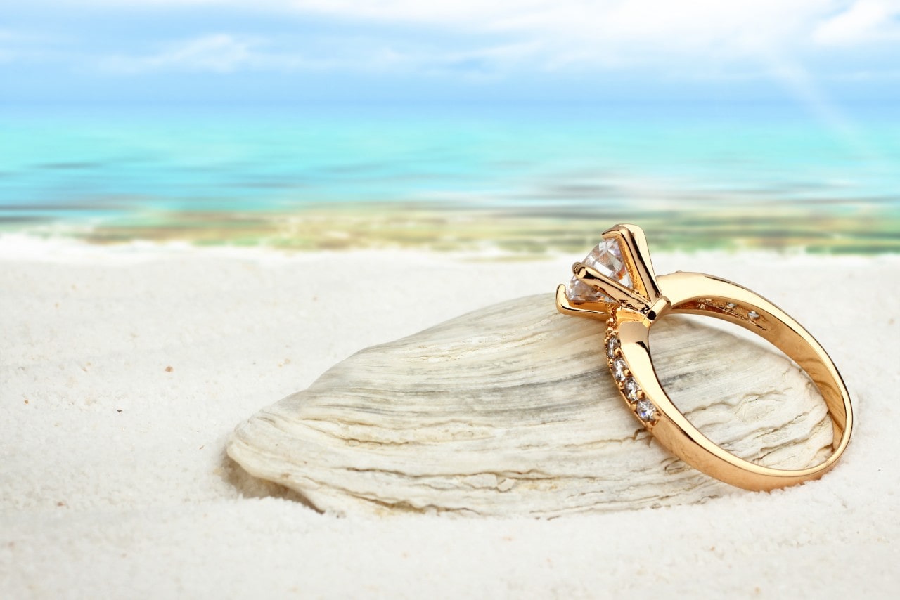 A gold engagement ring with a prong set center stone leaning against a shell on the beach
