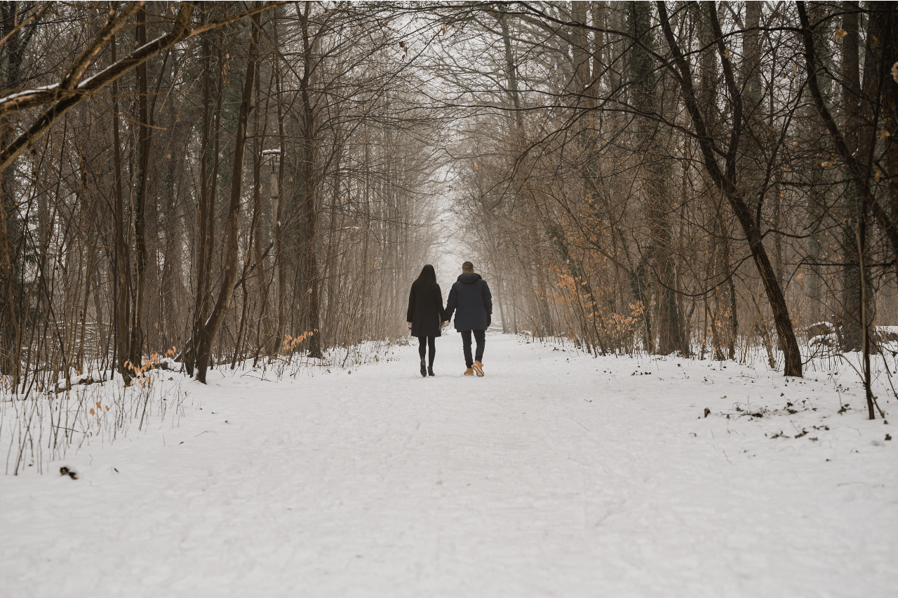 A couple walking down a snowy path in the woods and holding hands