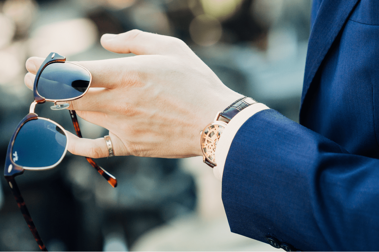 A man wearing a elaborate watch under his suit plays with his retro sunglasses