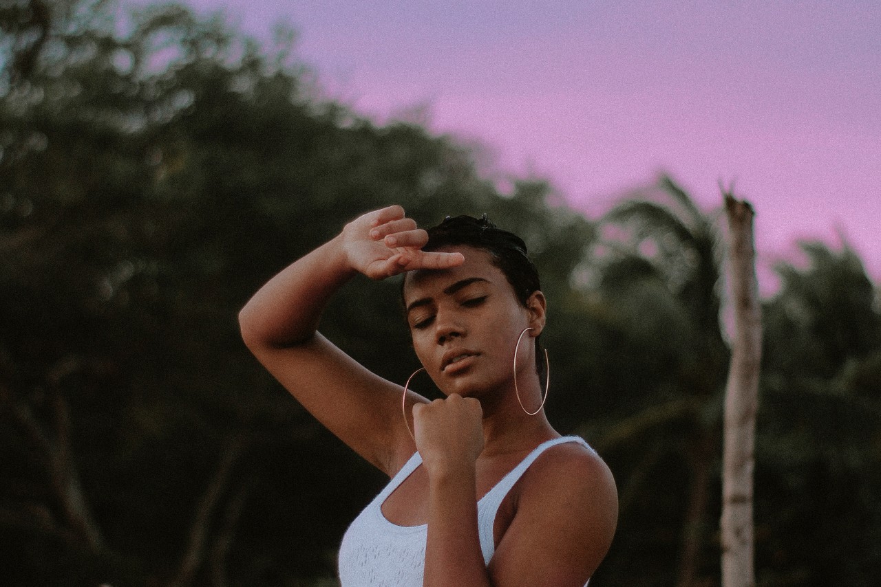 A woman wearing a white tank top and large gold hoop earrings shields her face on a beach