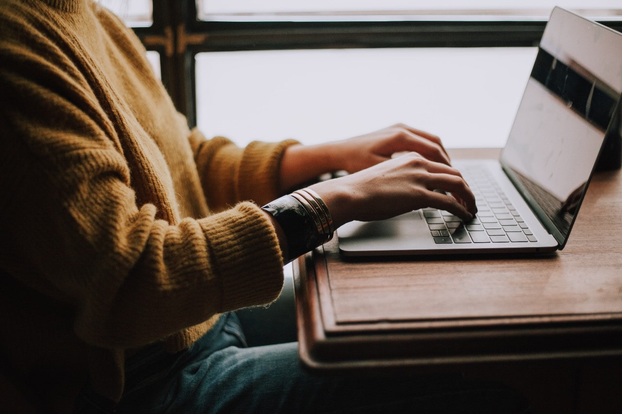 A person in a mustard yellow sweater typing on a computer and wearing a number of bracelets