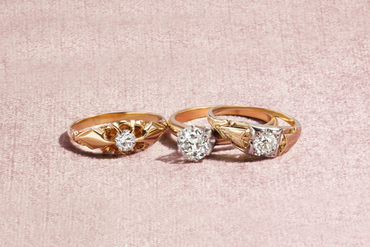 Three vintage diamond rings, each with a yellow gold setting and design, are displayed in a row against a soft, textured pink background