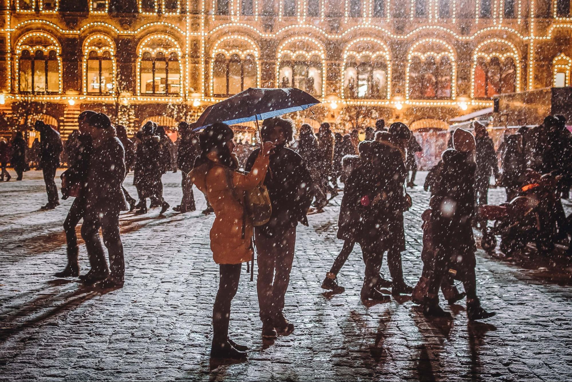 A young couple standing in front of a lavishly lit building as light snow falls.