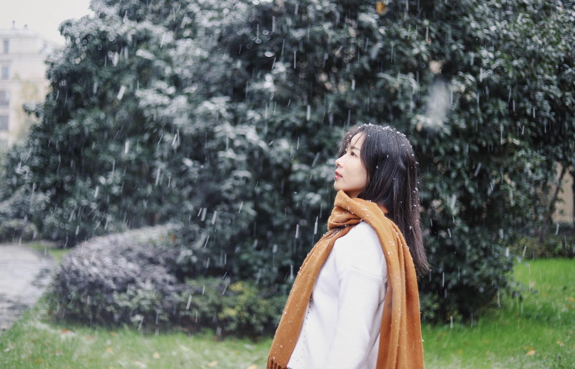 A warmly dressed young woman standing outside as snow lightly falls.