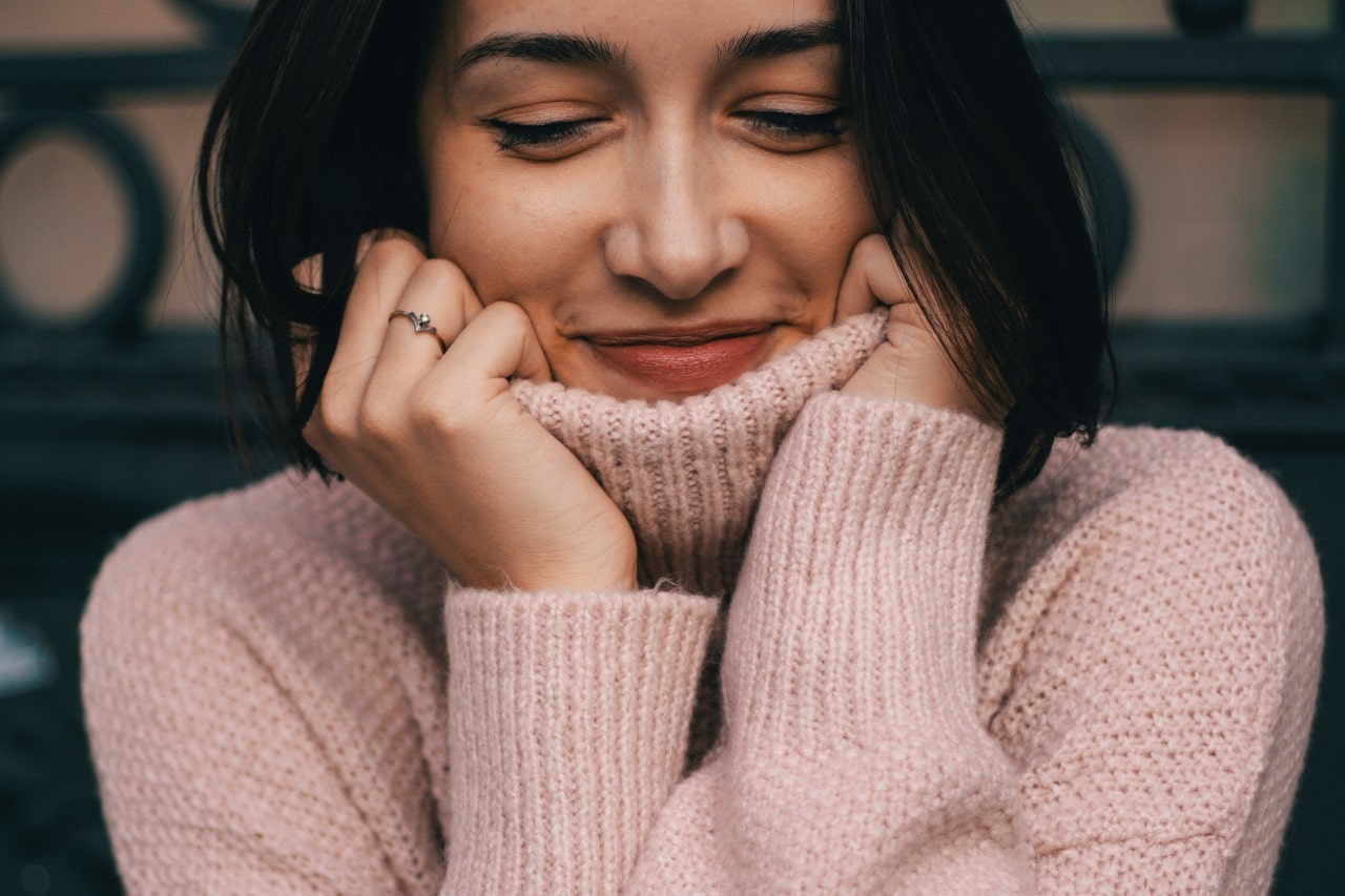 A close-up of a smiling woman in a comfortable sweater, a delicate engagement ring on her finger.