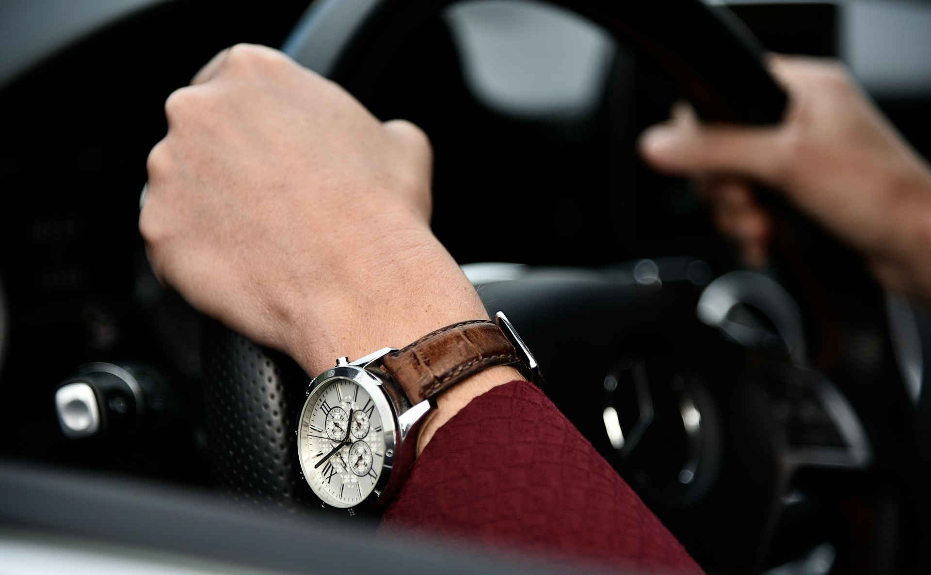 a pair of hands holding a steering wheel, one wrist adorned with a vintage-inspired watch