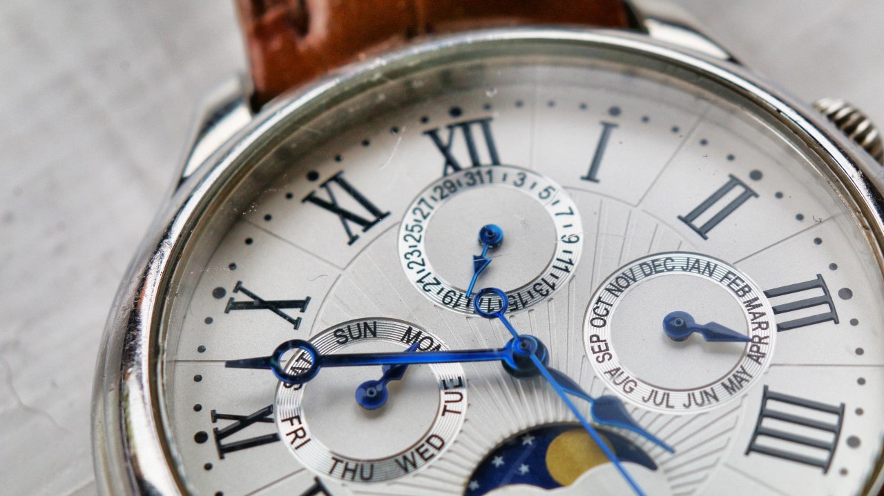 A close up of a watch dial with blue hands, roman numbers, and brown leather strap. A close up of a watch dial with blue hands, roman numbers, and brown leather strap.