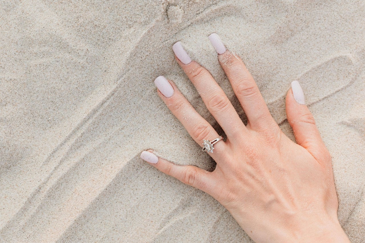 A woman’s hand in the sand adorned with a dainty engagement ring.