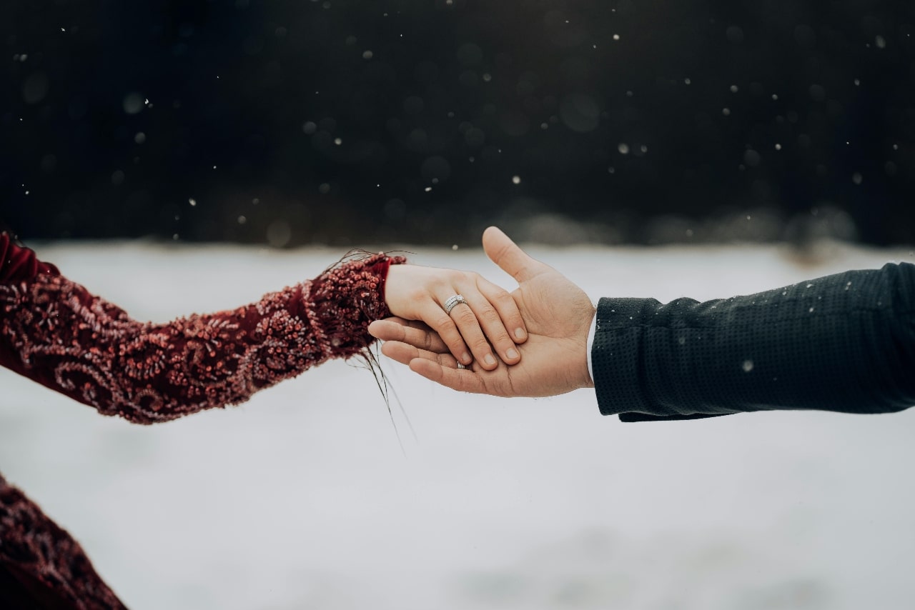 A couple holding hands, showcasing a diamond wedding band on the woman's hand against a snowy background