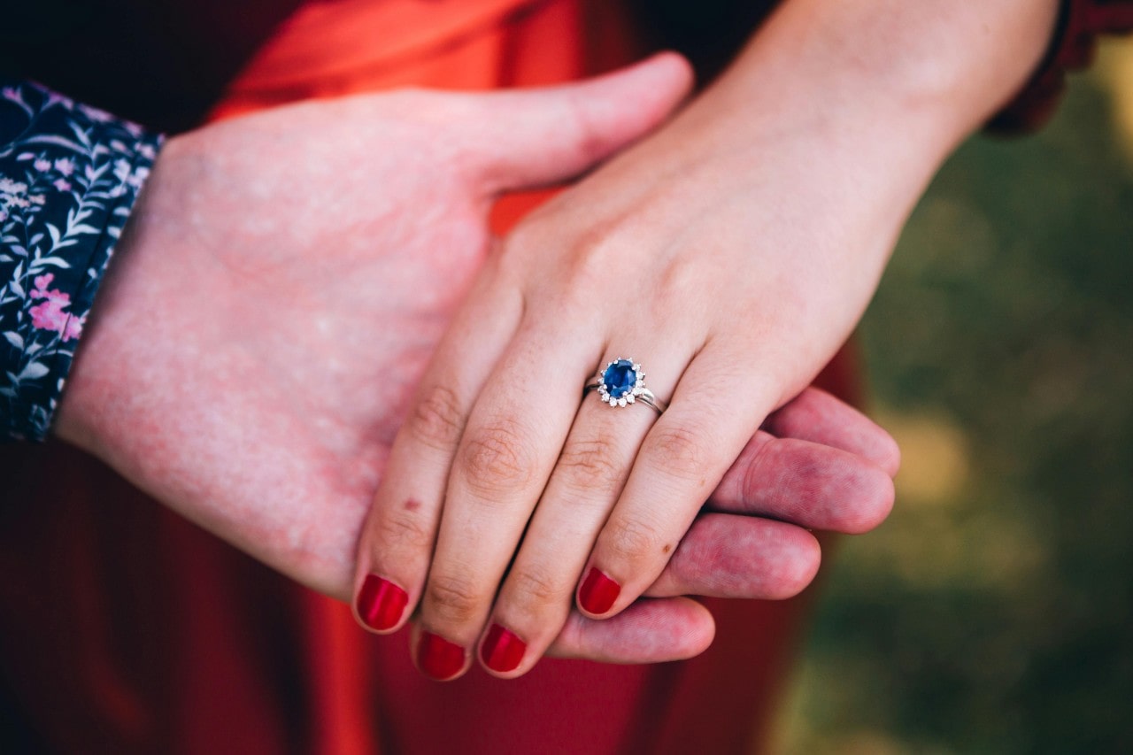 A man’s hand holding a woman’s, the woman wearing a sapphire engagement ring.